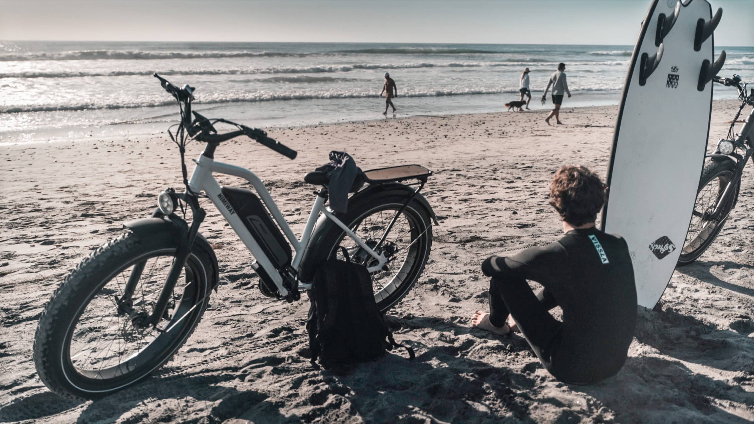 Himiway fat tire electric bike is on a beach with a surfer and a surfboard